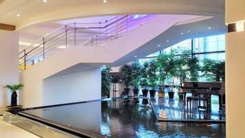 Un espacio interior amplio con un elemento acuático reflectante, plantas en macetas y un piano.