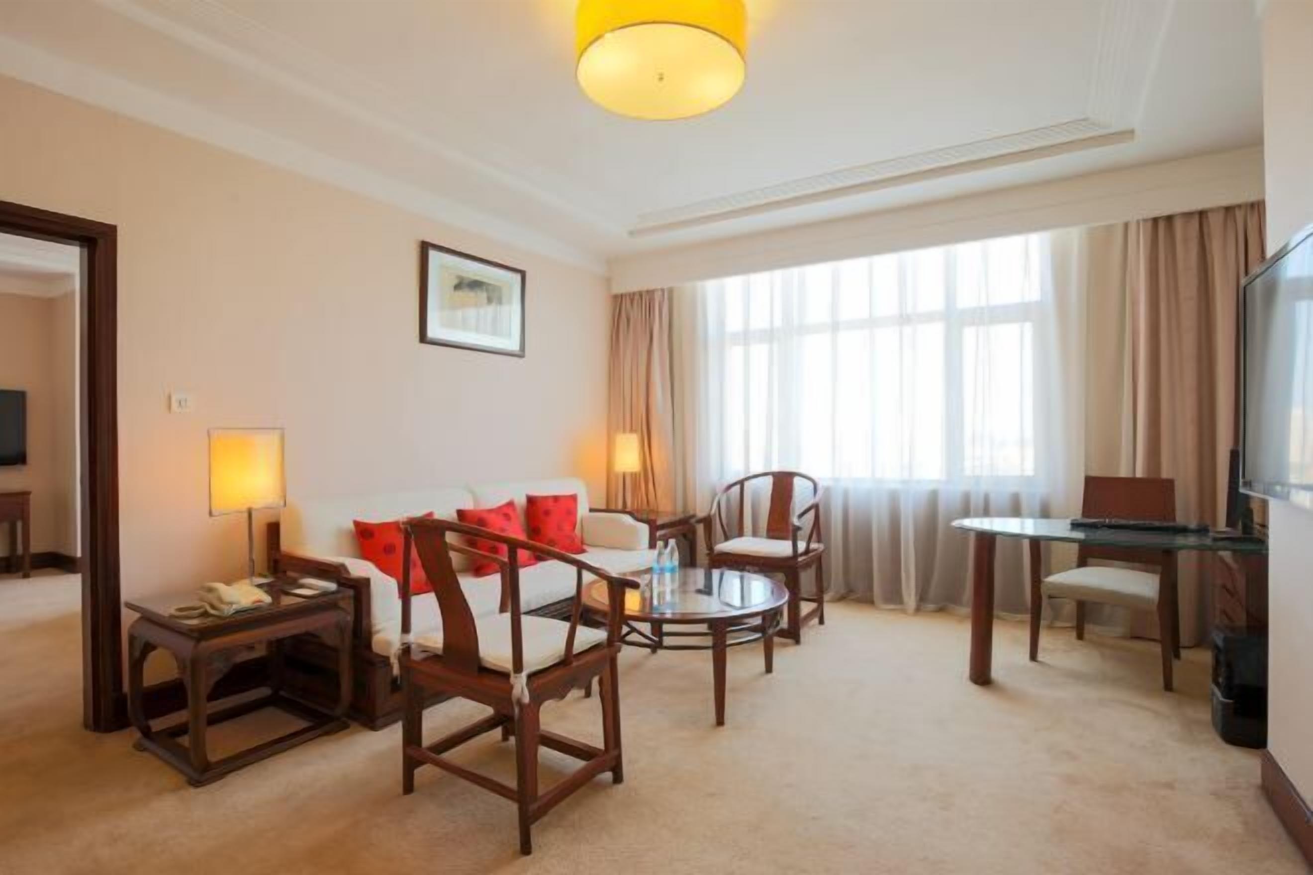 Habitación de hotel con sofá, dos sillones, una mesita, televisor y ventana grande con cortinas.