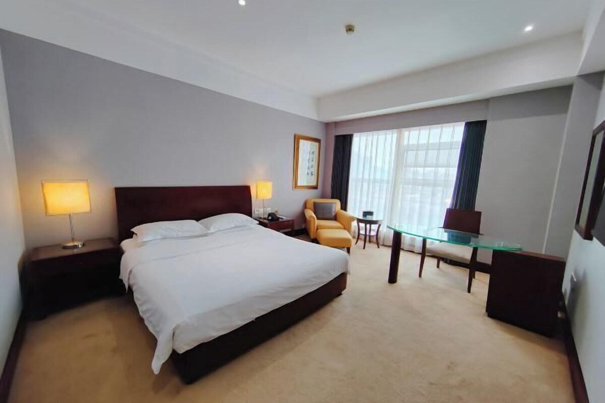 Une chambre d’hôtel avec un grand lit, deux tables de chevet avec des lampes, une petite table avec une chaise et une grande fenêtre avec des rideaux.