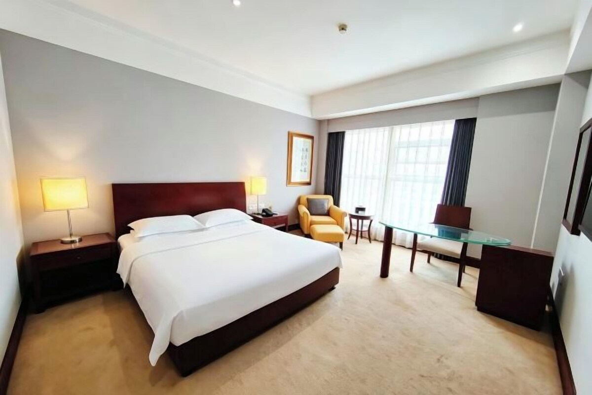 A hotel room with a large bed, bedside lamps, a desk, a chair, and a window with curtains.