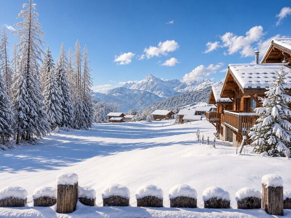 A snow-covered wooden lodge with a mountain backdrop, wooden fences, and snow-covered trees.