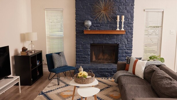 A living room with a stone fireplace, a blue chair, a gray sofa, a white table, and a patterned rug. - 2BR Ensuite | Near UT & The Domain (Austin)