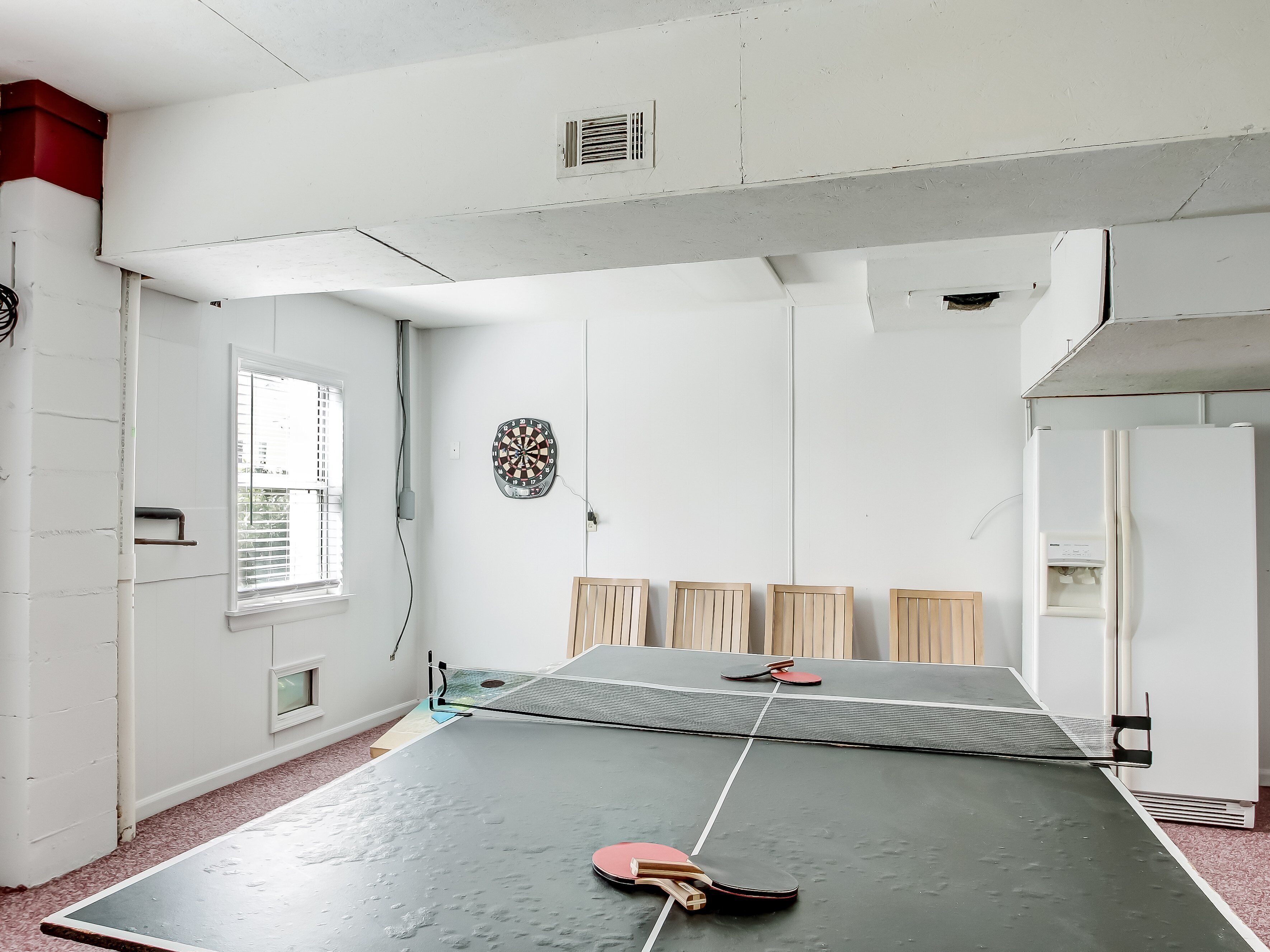 A ping pong table with paddles and a dartboard in a room with white walls and a window.