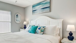 Un dormitorio con cama blanca, ventilador de techo, un cuadro del océano, espejo redondo y mesita de noche con lámpara.