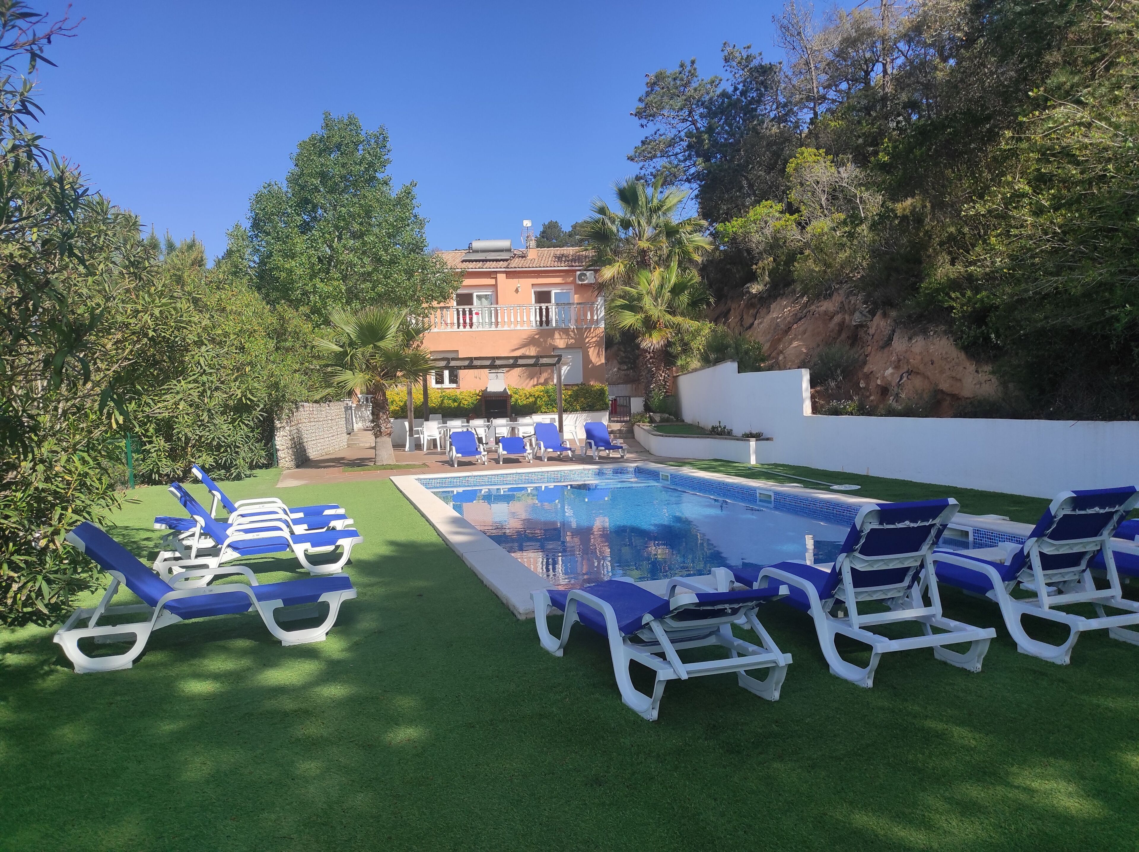 Una piscina con sillones de jardín azules y blancos alrededor, un edificio al fondo y vegetación circundante.