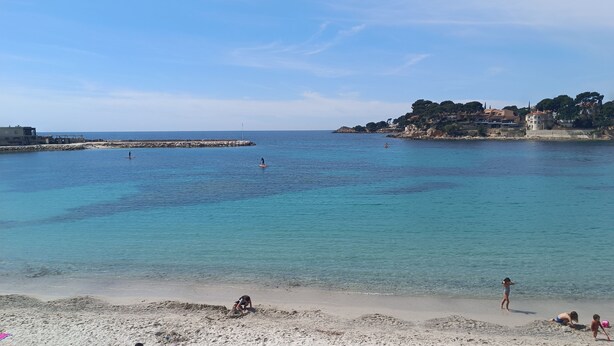Ein Strand mit klarem türkisblauem Wasser, einem sandigen Ufer und Gebäuden im Hintergrund.