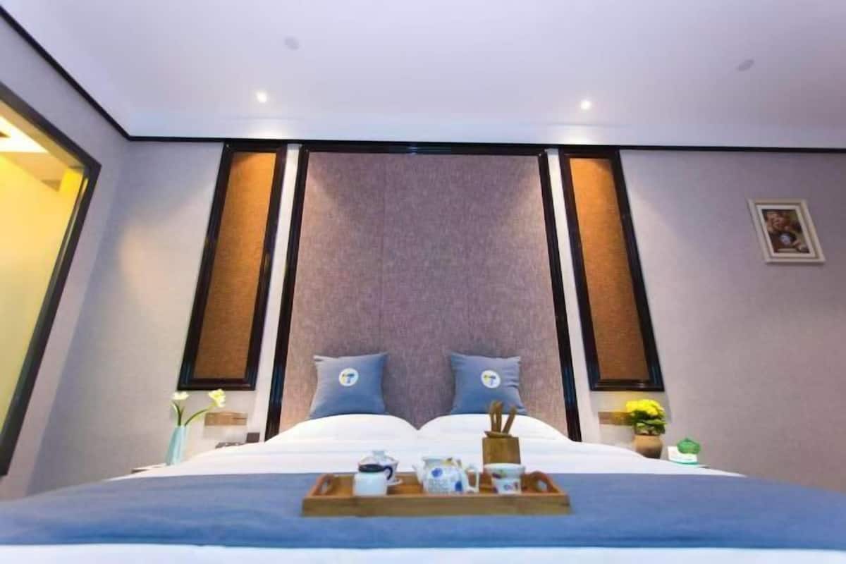 Una habitación de hotel moderna con una cama grande, dos almohadas, una bandeja con té y tazas, y una pequeña planta en la mesita de noche.