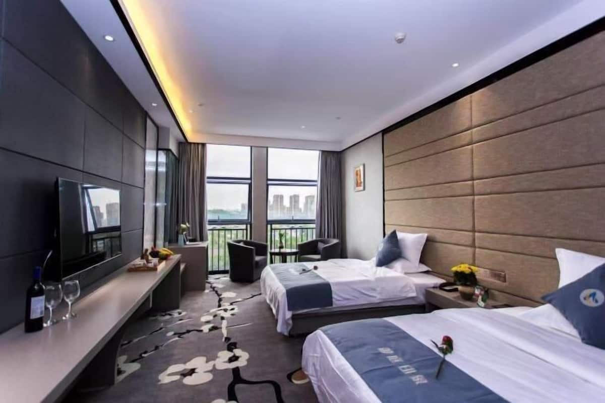 Habitación de hotel con televisor grande, dos camas, escritorio y balcón con vistas a la ciudad.