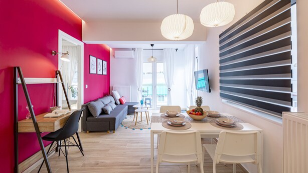 A modern living room with a dining area, featuring a bright red wall, a grey sofa, a wooden dining table, and a large window with blinds.