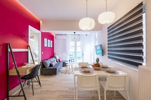 A modern living room with a dining area, featuring a bright red wall, a grey sofa, a wooden dining table, and a large window with blinds.