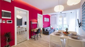A modern living room with a red accent wall, a grey sofa, a dining table with a fruit bowl, and a white ceiling fan.