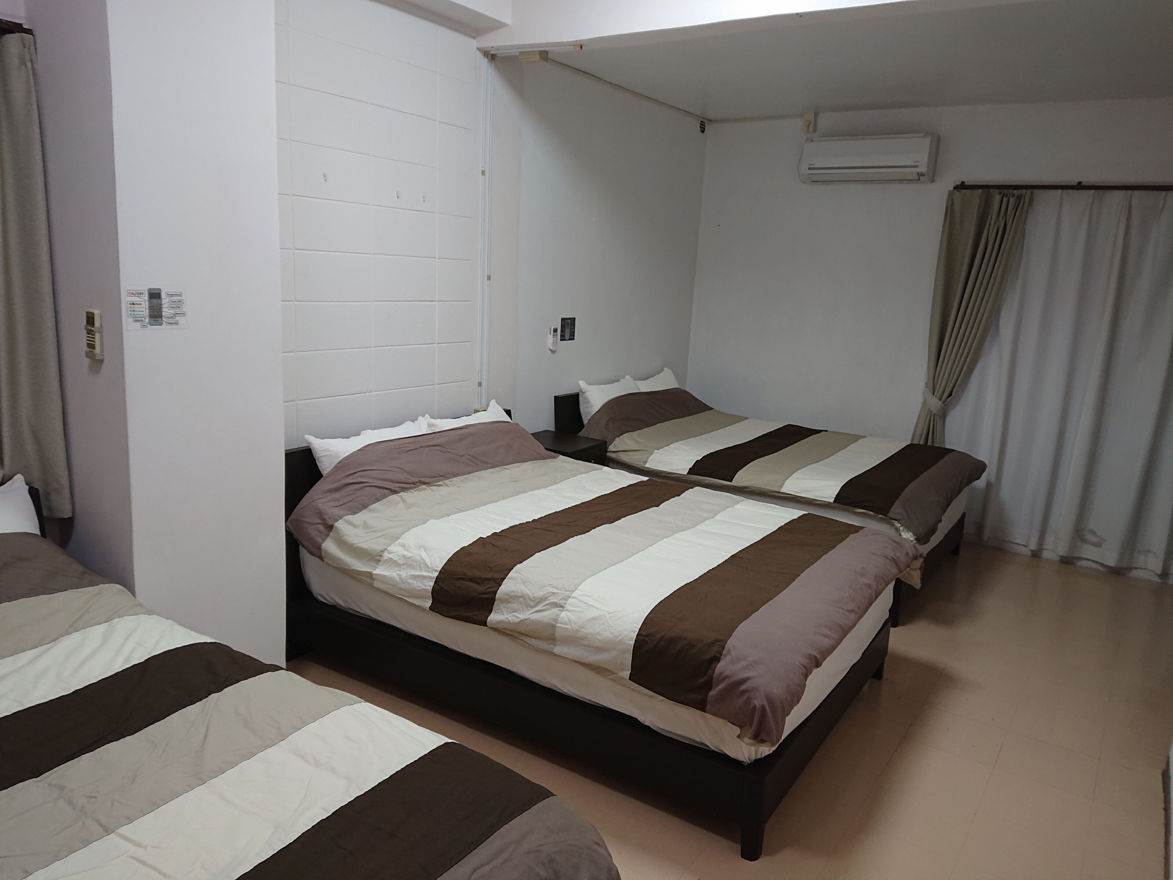 Une chambre d’hôtel avec deux lits, un mur blanc, un appareil de climatisation et un rideau.