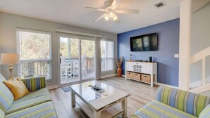 A living room with a ceiling fan, a TV mounted on the wall, a sofa, a coffee table, and a sliding glass door leading to an outdoor area. - PRIVATE BEACH - Gulf Peeks - Newly renovated - Bikes included - 7 Palms (Panama City Beach)