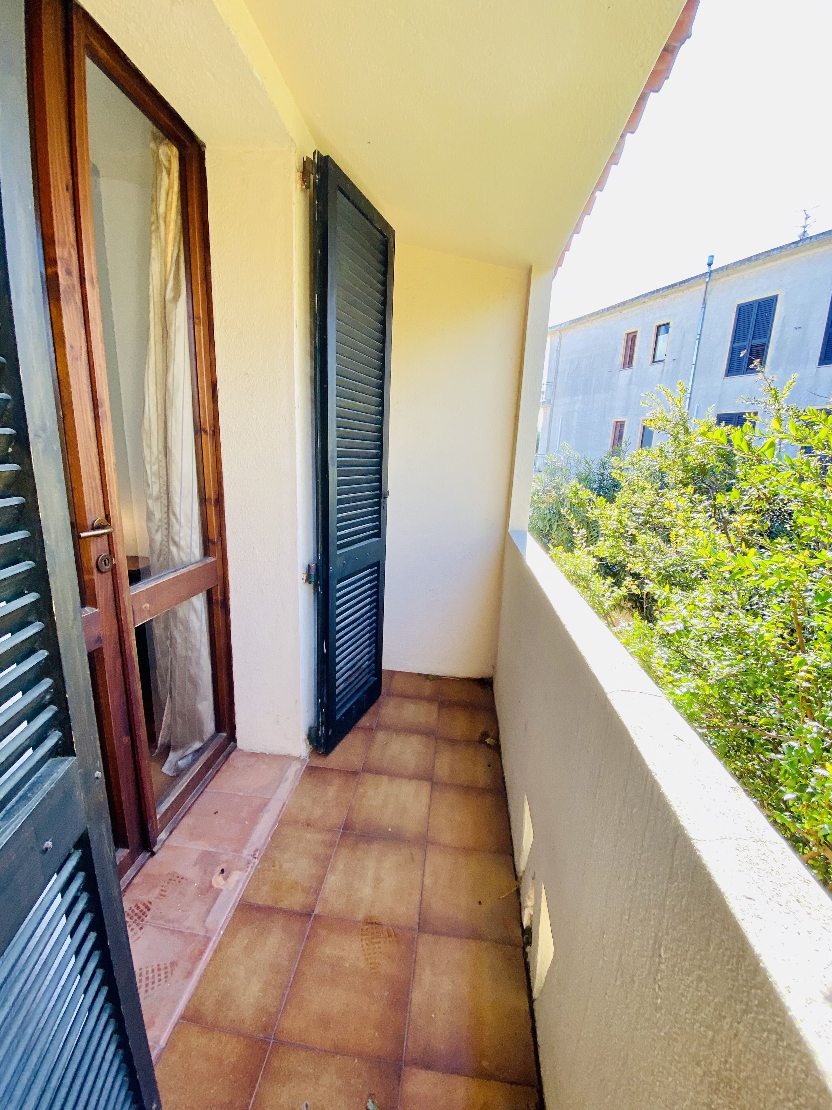 A narrow balcony with a tiled floor, a wooden door with a glass panel, and a black shutter door.