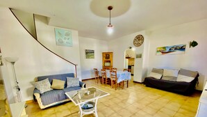 A living room with a sofa, a coffee table, a dining table with chairs, a kitchen area, and a staircase. - L'avru Di La Canna La Cinta (San Teodoro)