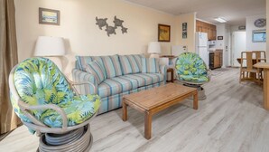 Ein Wohnzimmer mit einem gestreiften Sofa, zwei Sesseln, einem Holztisch und einer sichtbaren Küchenzeile im Hintergrund.