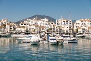 A marina with boats docked, surrounded by white buildings with red-tiled roofs, and a mountain in the background.