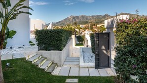 Un espacio exterior moderno con una escalera de hormigón que lleva a una entrada protegida por una verja, rodeado de vegetación y paredes blancas.