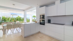 Una cocina moderna con muebles blancos, una zona de comedor con mesa de vidrio y vistas al exterior.