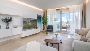 A modern living room with a flat-screen TV, a coffee table, and a sofa. There is a potted plant, a chair, and a view of the outdoors through a glass door. - NEWLY BUILT Top Modern Penthouse in Puerto Banús (Puerto Banús)