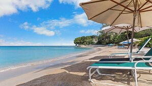 A beach with clear blue water, a sandy shore, and lounge chairs under umbrellas. - 1 bedroom Beachfront Condo, small pool, gorgeous sea view (Paynes Bay)