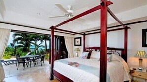 A four-poster bed with a white quilt, a wooden nightstand with a lamp, and a view of palm trees through a glass door. - 1 bedroom Beachfront Condo, small pool, gorgeous sea view (Paynes Bay)