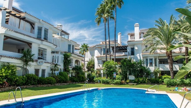Une piscine entourée de bâtiments blancs dotés de balcons, de palmiers et surplombant un ciel bleu azur.