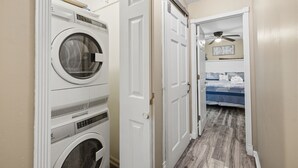 Ein Waschraum mit gestapelten Waschmaschinen und Trocknern, eine Tür zu einem Schlafzimmer mit Bett und Deckenventilator.