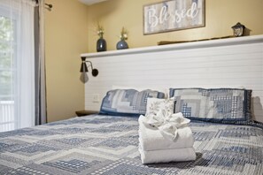 A neatly made bed with a patterned bedspread, a folded towel, and a wall with a 'Blessed' sign.