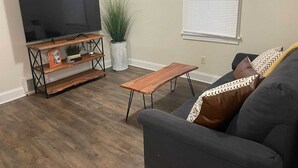 Ein Wohnzimmer mit Flachbildfernseher auf einem Holzboden, einem Blumentopf, einer Bank und einem Sofa mit gemusterten Kissen.