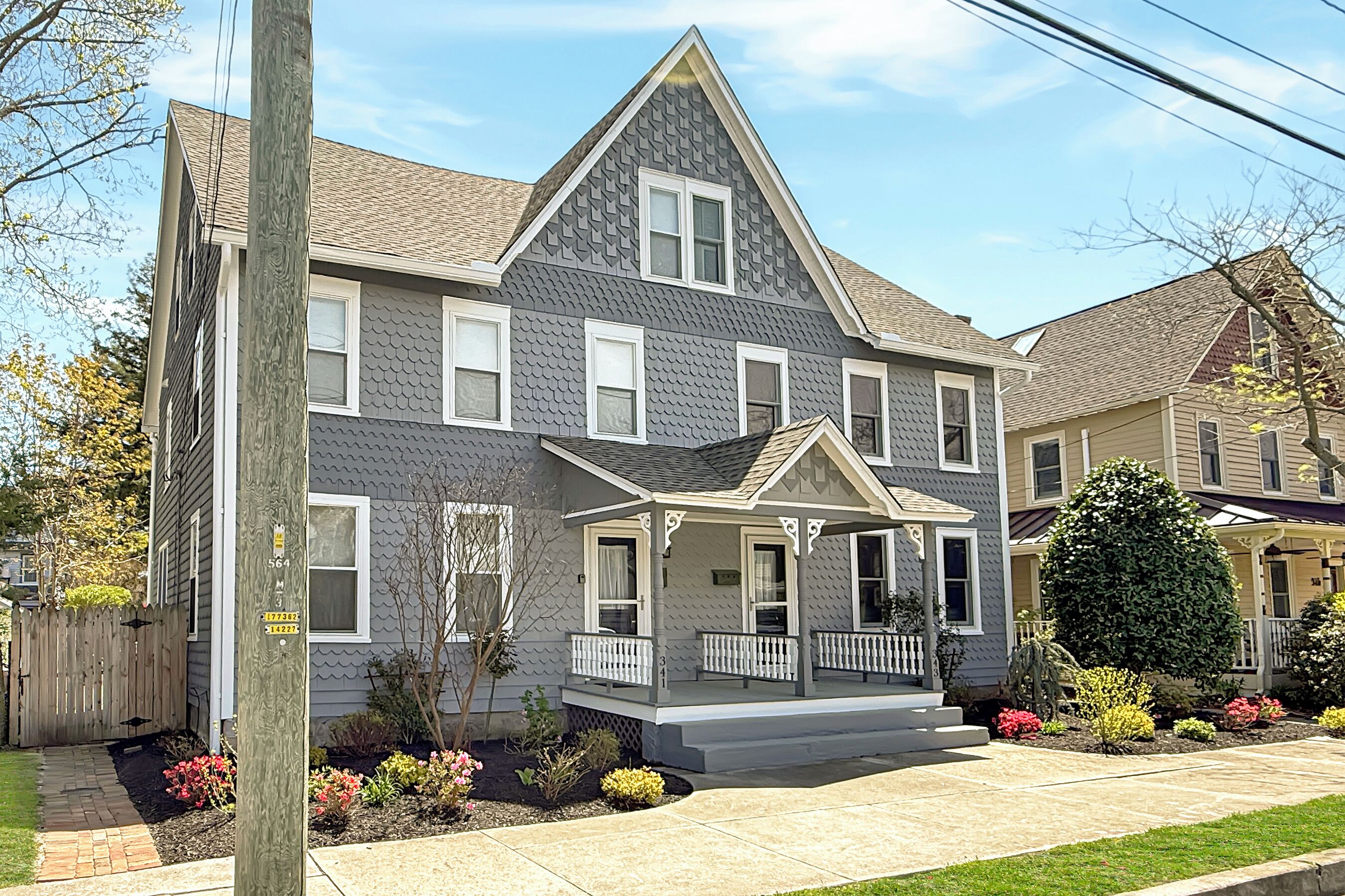 A two-story house with a gabled roof, a front porch, and a small front yard with various plants and flowers.