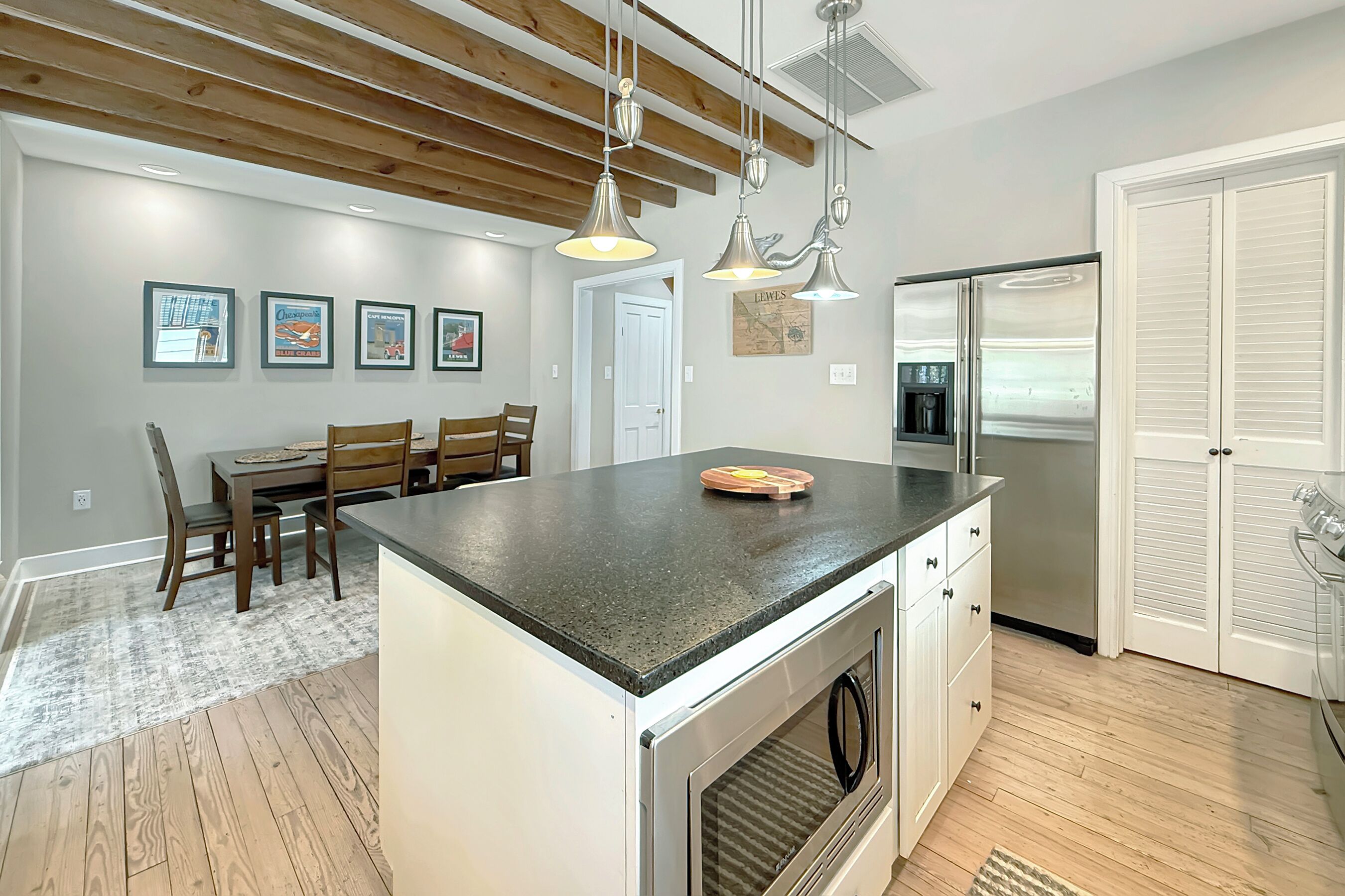 A kitchen with a central island, wooden ceiling beams, pendant lights, a refrigerator, and a dining area with chairs and a table.