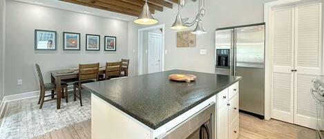 A kitchen with a central island, wooden ceiling beams, pendant lights, a refrigerator, and a dining area with chairs and a table.