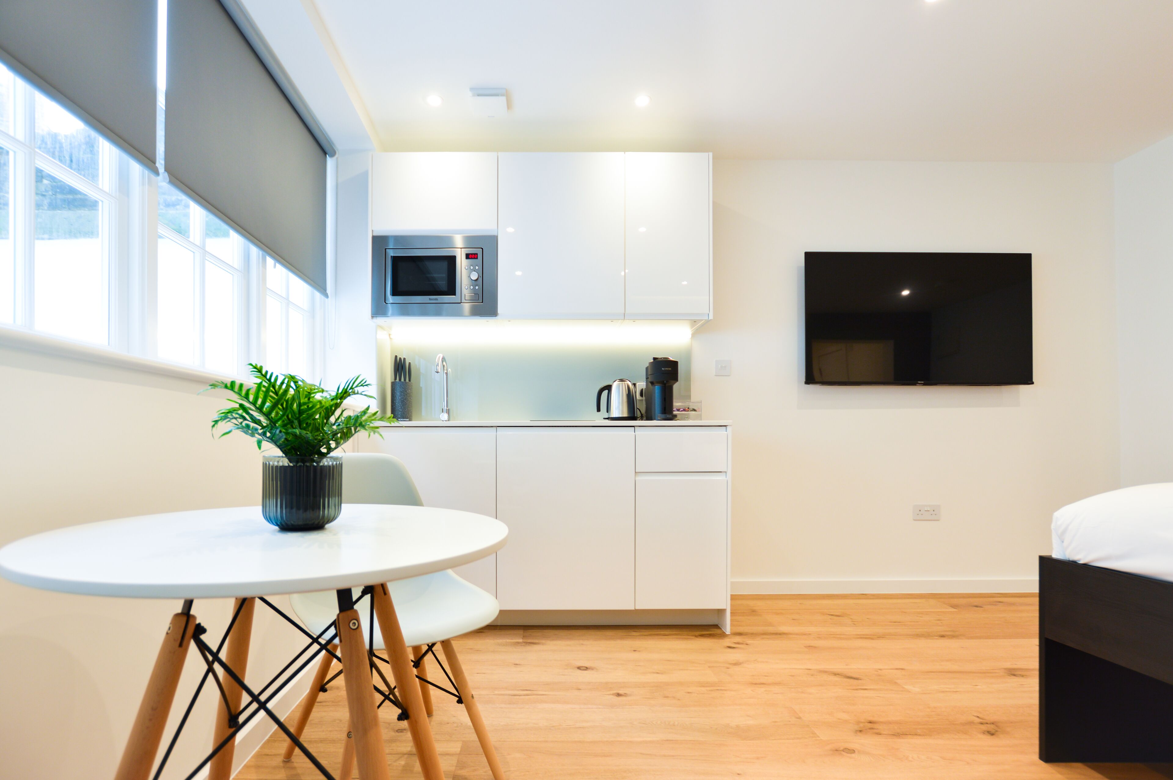 A modern kitchen with white cabinets, a microwave, and a coffee maker. There is a dining table with chairs and a potted plant. A TV is mounted on the wall.