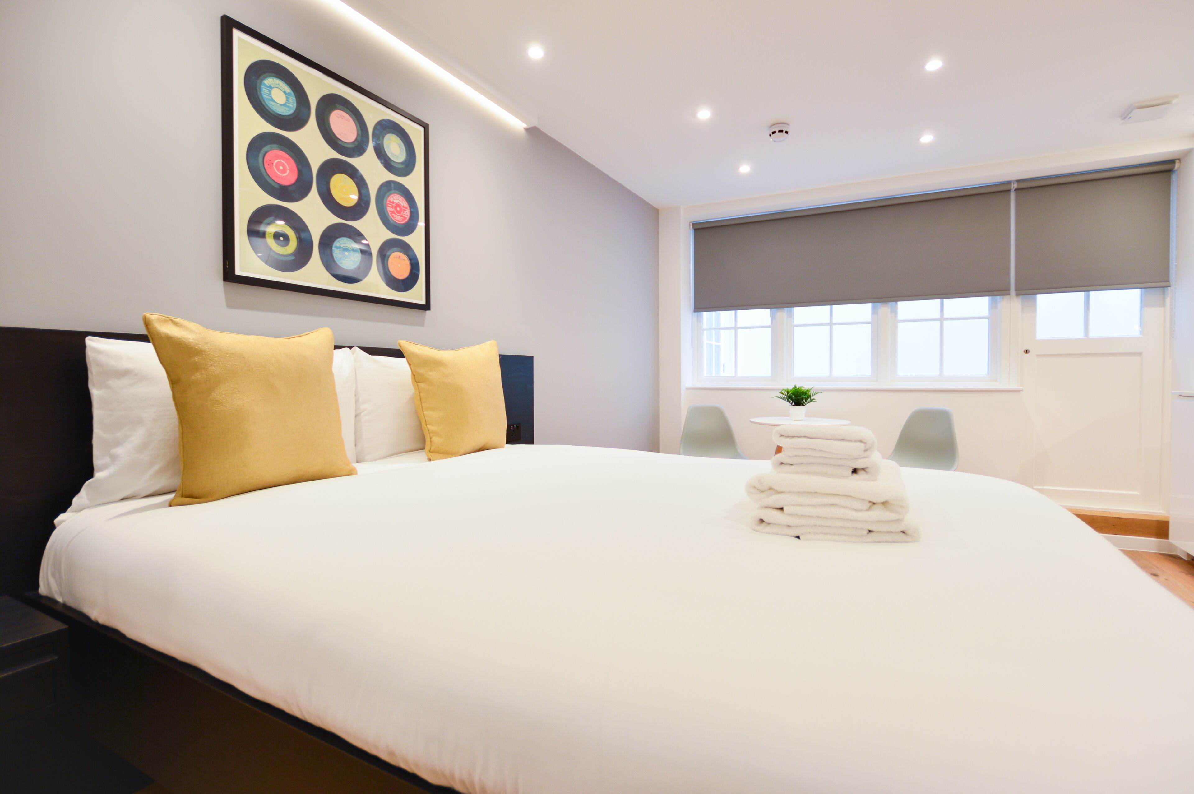 A modern hotel room with a large bed, a headboard, a window with blinds, a painting on the wall, and a small plant on the bed.