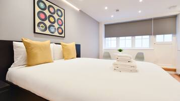 A modern hotel room with a large bed, a headboard, a window with blinds, a painting on the wall, and a small plant on the bed.