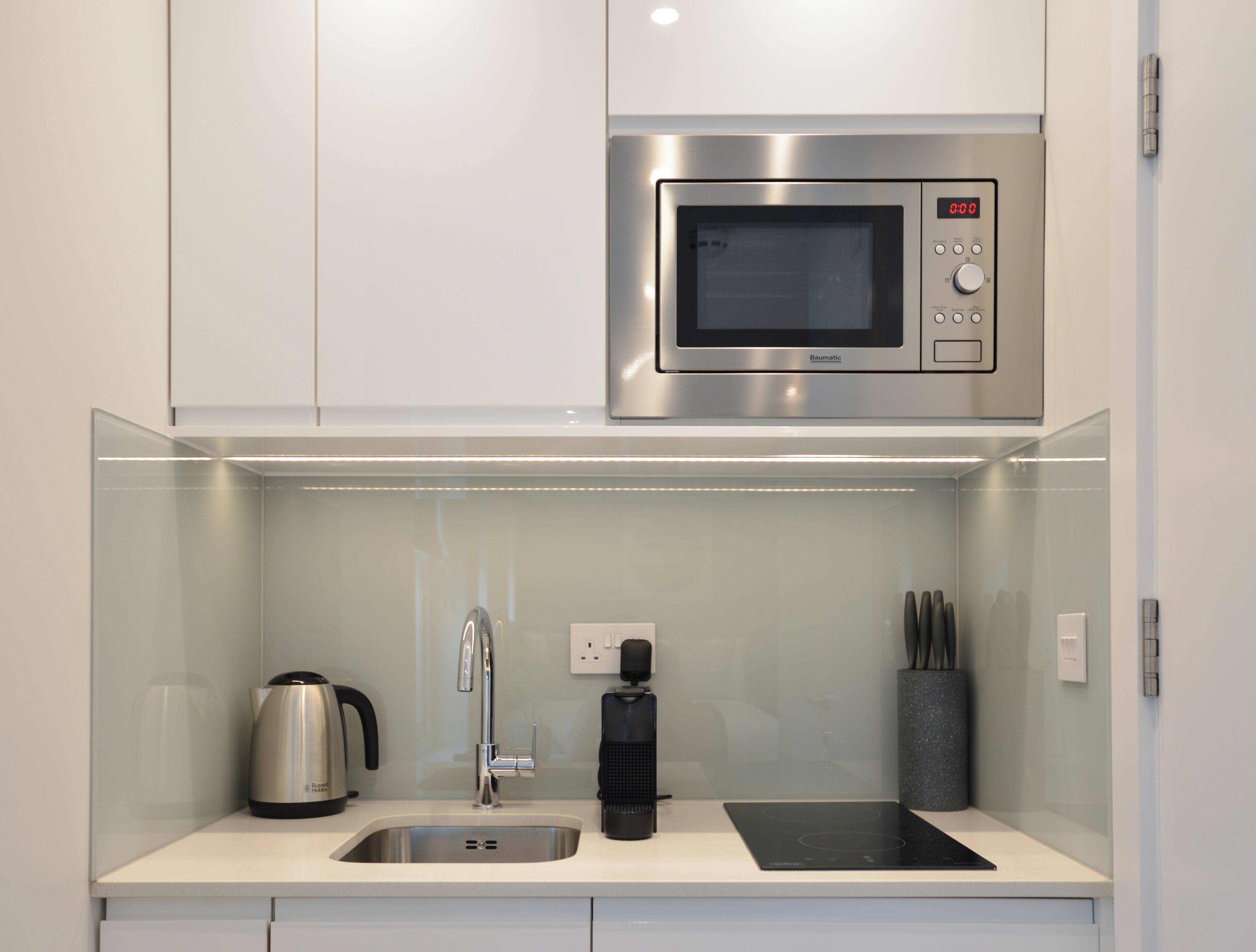 A compact kitchen with a stainless steel microwave, a kettle, a coffee maker, and a black induction cooktop.
