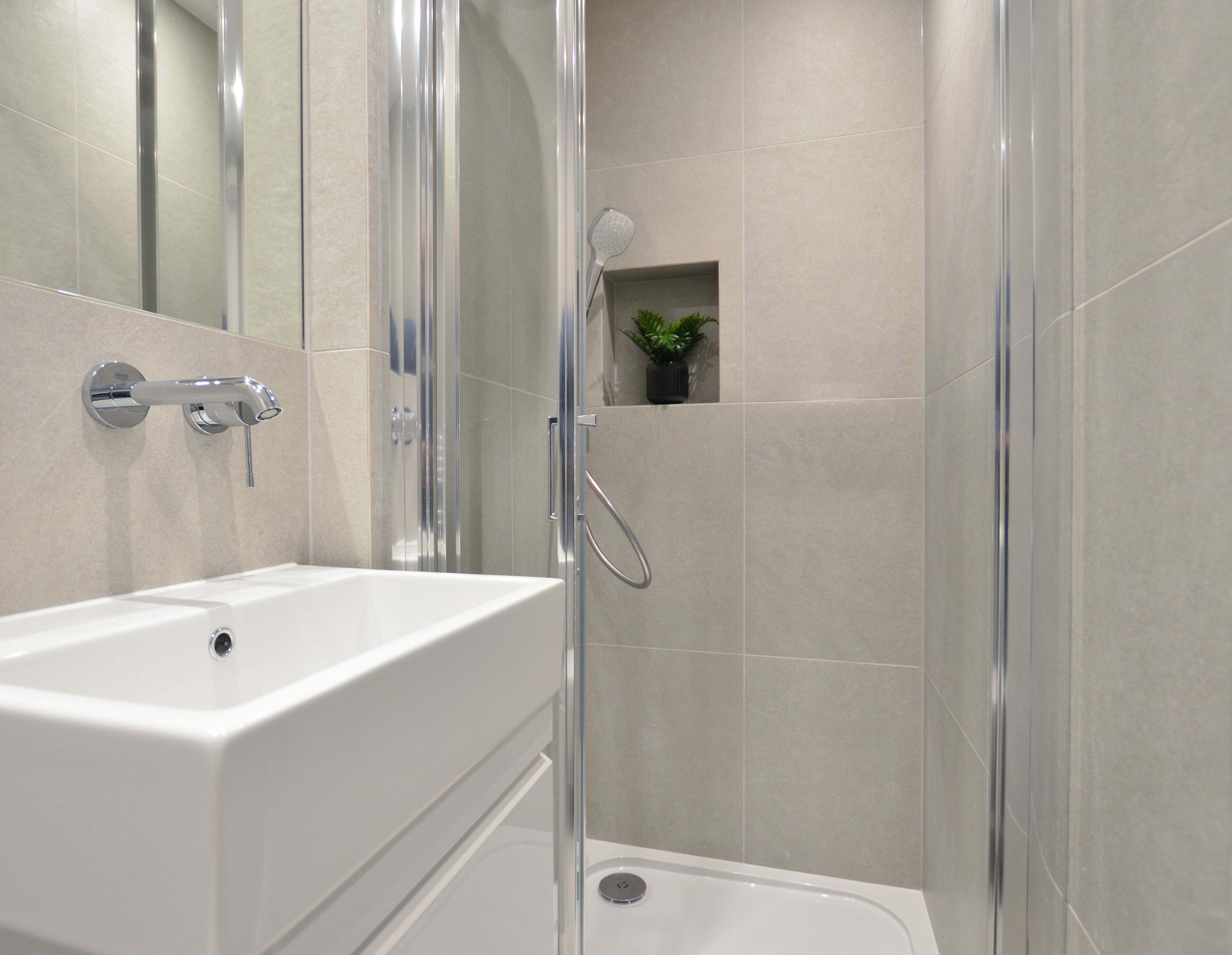 A modern bathroom with a white sink, a glass-enclosed shower, and a small potted plant on the wall.