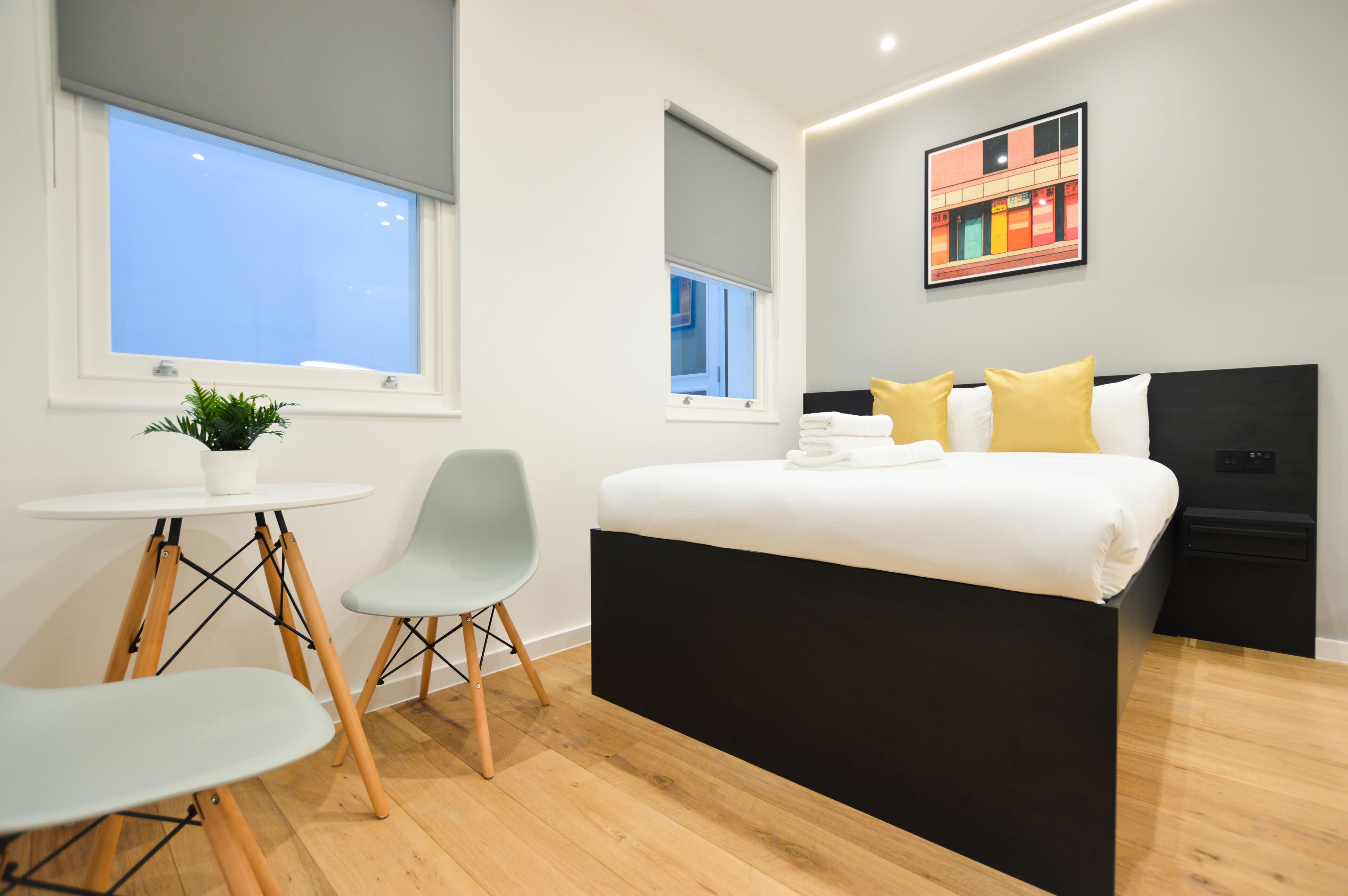 Un dormitorio moderno con una cama grande, una mesita pequeña, dos sillas, una planta en maceta y un televisor montado en la pared.