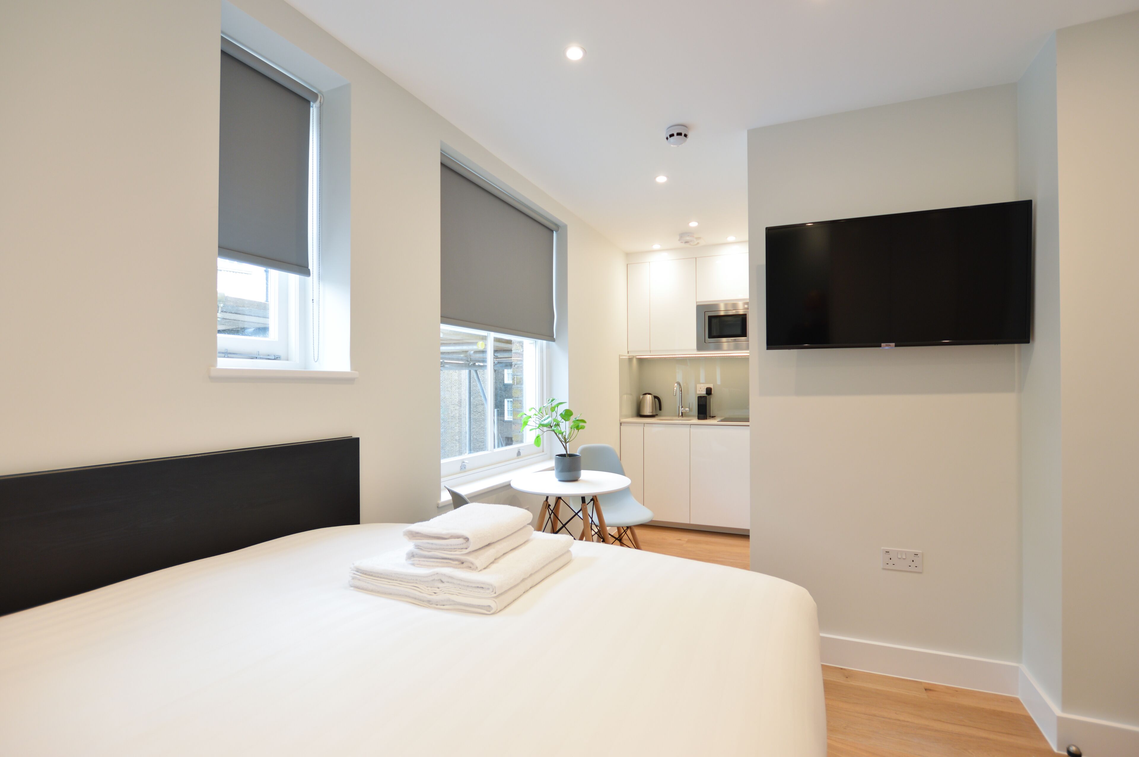 Ein modernes Hotelzimmer mit einem großen Bett, einem kleinen Esstisch, einer Zimmerpflanze und einer Küchenzeile mit Fernseher.
