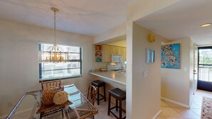 A compact kitchen with a microwave, a coffee maker, and a window with blinds. There is a dining area with a glass table and chairs, and a decorative wall hanging. - Prime Location I Free Beach Access Directly Across The Street!!! (Marco Island)