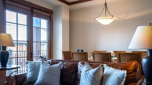 Een gezellige woonkamer met een bruine leren bank, een ronde tafel met een lamp en een eethoek met rieten stoelen.