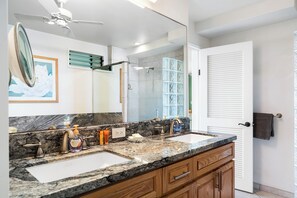 A bathroom with a large mirror, two sinks, wooden cabinets, a ceiling fan, and a glass shower enclosure. - KKSR#4102 DIRECT OCEANFRONT, INCREDIBLE GROUND FLOOR UNIT, REMODELED!, AC!!! (Kailua Kona)