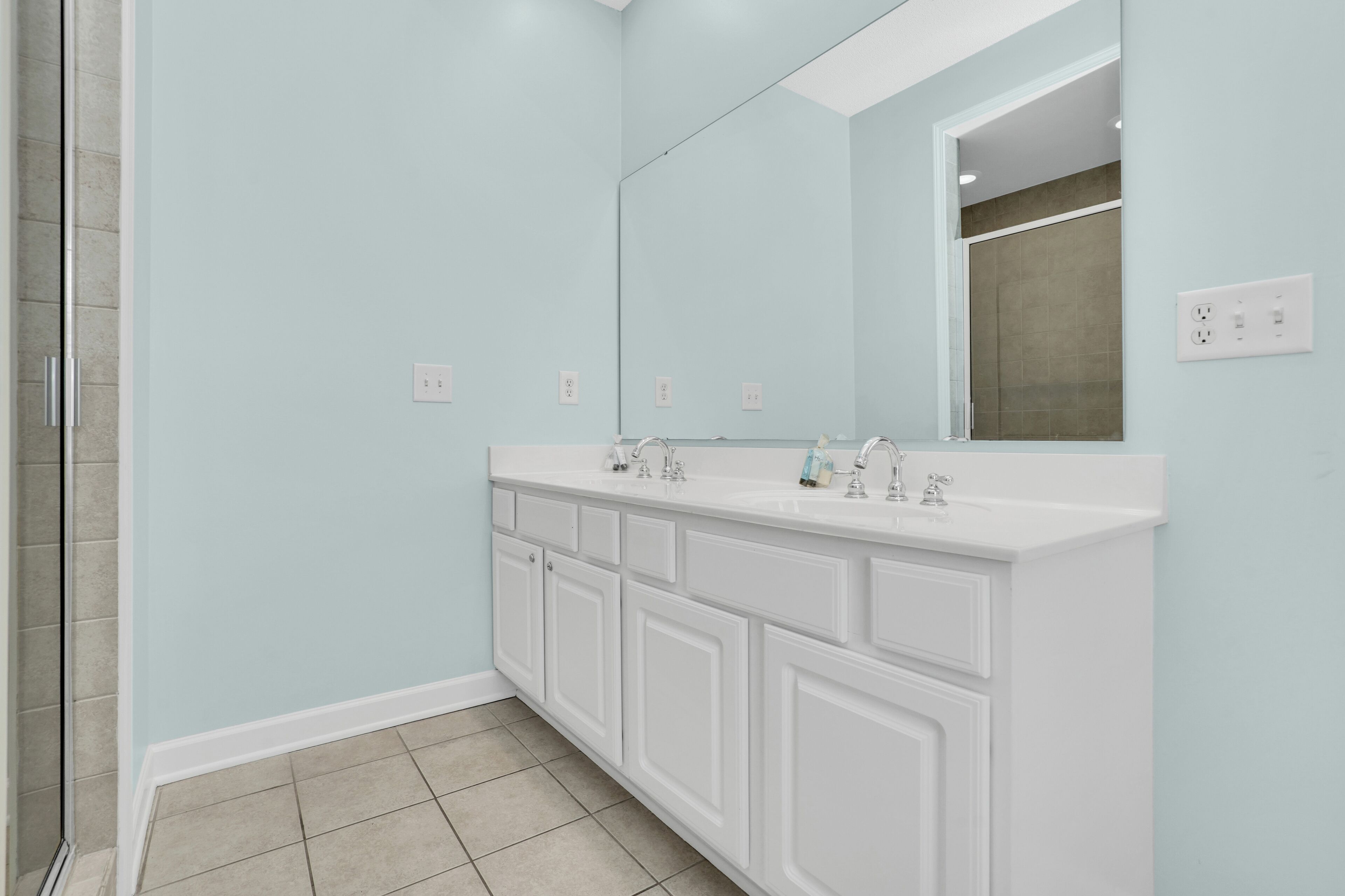 A bathroom with a light blue wall, white vanity with multiple drawers, a large mirror, and a shower area.
