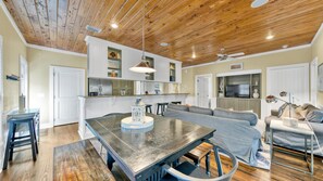 A spacious living area with a large wooden dining table, a grey sofa, and a television in the background.