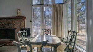 Un salon chaleureux avec une cheminée en pierre, une table à manger avec des chaises et de grandes fenêtres donnant sur un paysage arboré et une terrasse.