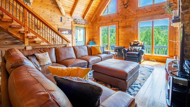 A wooden cabin interior with a large leather sofa, a fireplace, and a staircase.