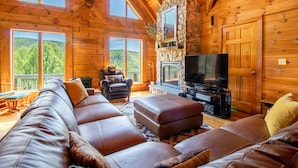 A log cabin interior with a large leather sofa, a stone fireplace, a television, and a view of the forest through large windows.