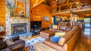 A log cabin interior with a stone fireplace, leather sofas, a television, and a kitchen area.