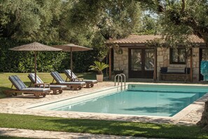 Ein Swimmingpool mit Liegestühlen und Sonnenschirmen, umgeben von Grünpflanzen und einem Steinhaus.